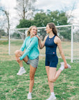MOVE Sport Apparel women’s Biker Shorts: Woman wearing Maritime Blue biker shorts during a standing quadriceps stretch on a grass field.