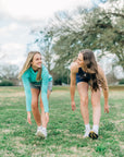 MOVE Sport Apparel women’s Biker Shorts: Woman wearing Maritime Blue biker shorts while stretching forward on a grass field.