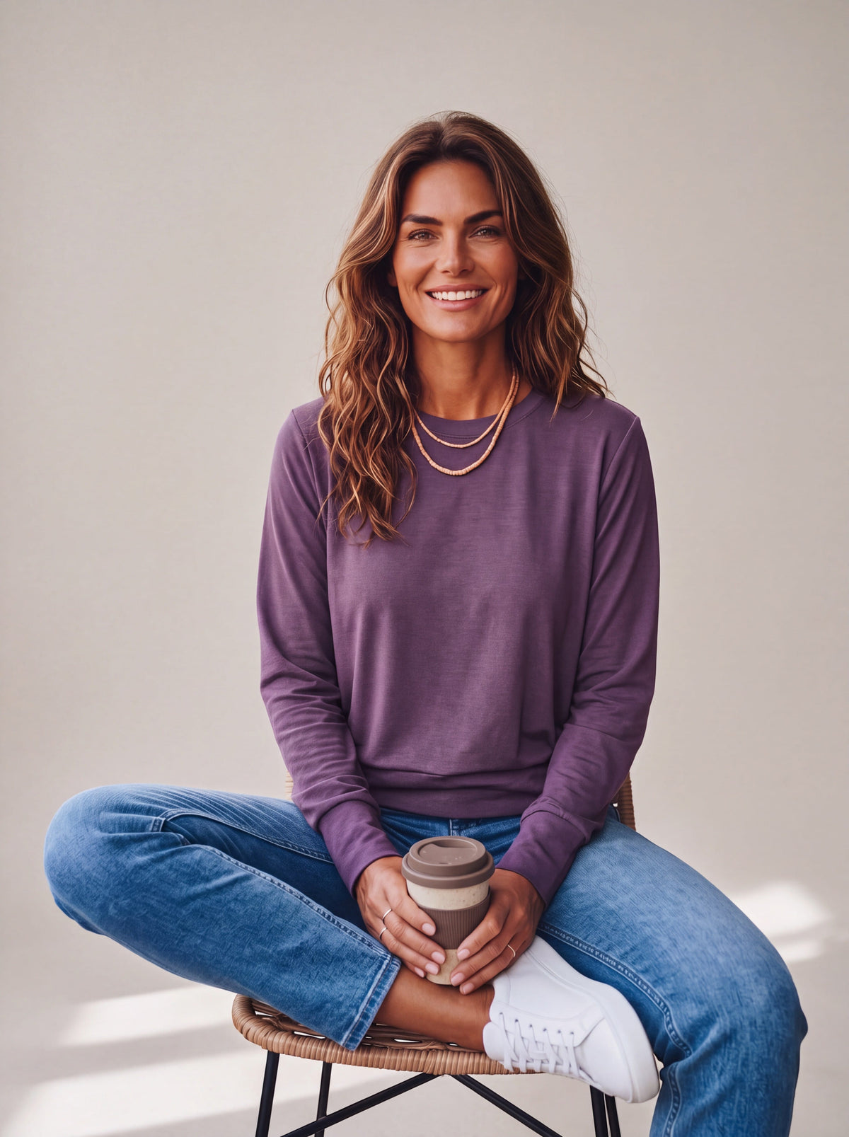 Woman seated in a neutral studio wearing the Gothic Grape MOVE Sport Apparel Women’s Long Sleeve Crewneck Pullover with blue jeans and white trainers, showing its natural fabric folds, crew neckline and ribbed cuffs.