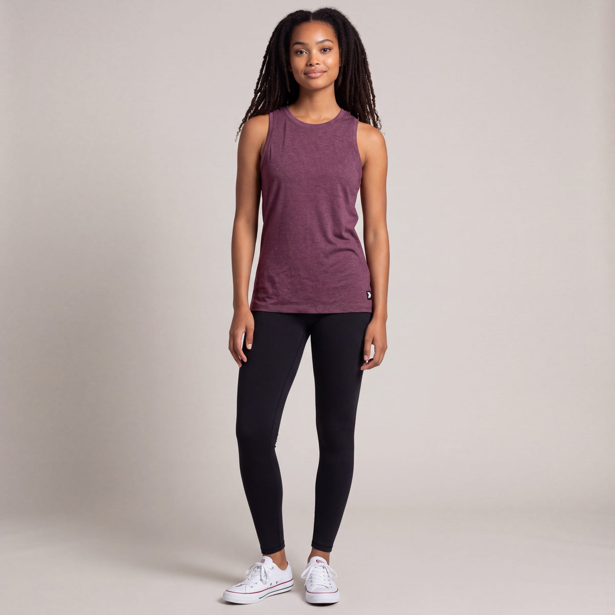 Full-body studio view of a woman wearing MOVE Sport Apparel women’s Essential High Neck Tank Top in Heather Fig with black leggings and white trainers; original small black-and-white hem tab retained.