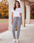 Woman wearing grey MOVE Sport Apparel Women’s Essential Joggers with a white tee and trainers beside an autumn running track and limestone pavilion, showing the original pocket tab and silver 3M reflective thigh embellishment.