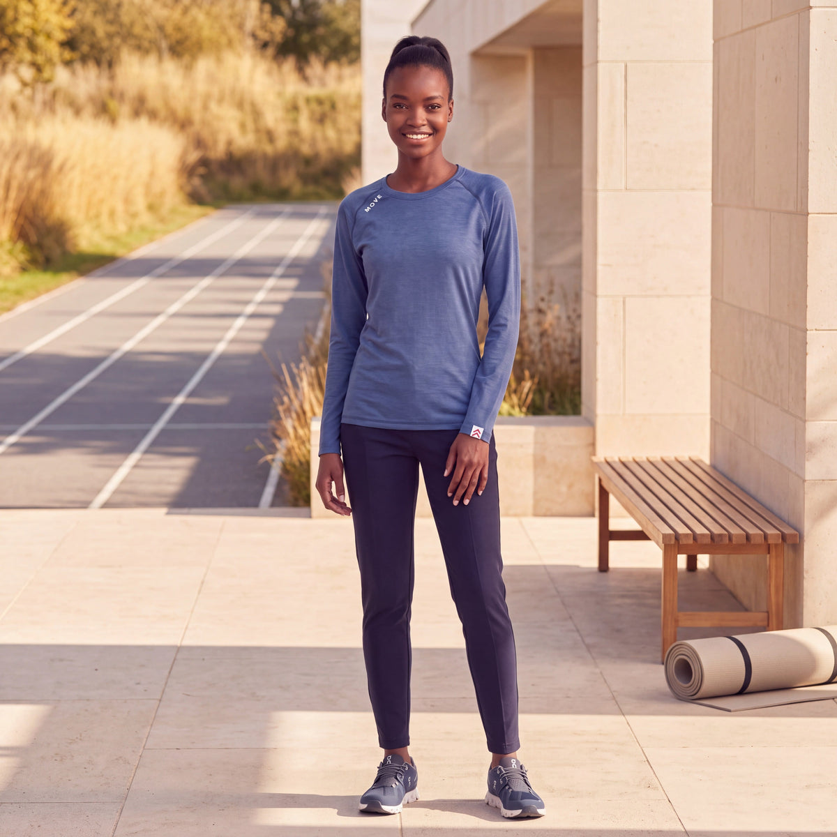 Woman wearing the heathered Surf the Web blue MOVE Sport Apparel Women’s Essential Long Sleeve at an autumn running-track recovery terrace; original white shoulder lettering, red-and-white cuff tab and natural hem retained.