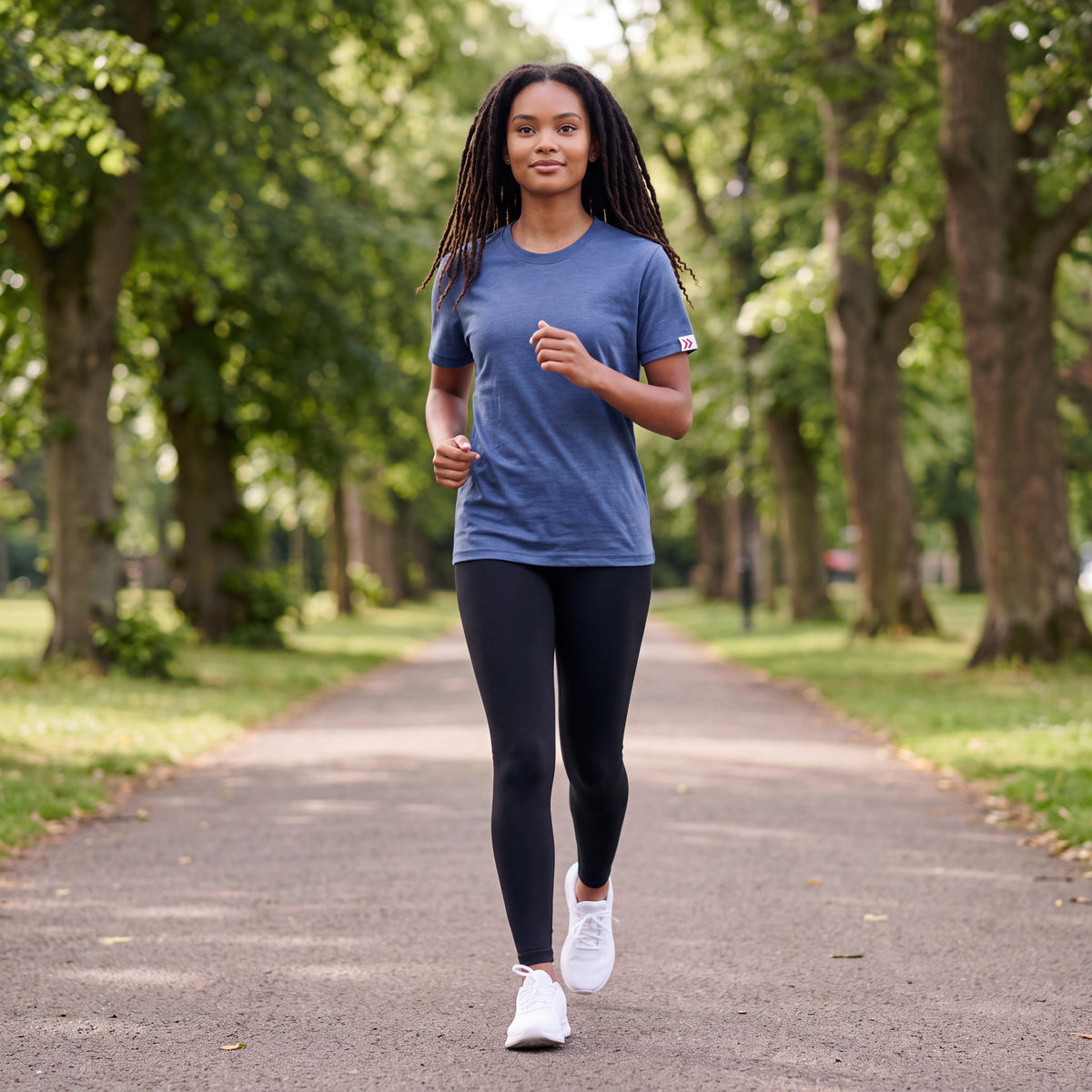 Woman jogging on a tree-lined park path in the Pageant blue MOVE Sport Apparel women’s Essential Short Sleeve T-shirt with black leggings and white running shoes.