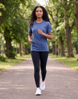 Woman jogging on a tree-lined park path in the Pageant blue MOVE Sport Apparel women’s Essential Short Sleeve T-shirt with black leggings and white running shoes.