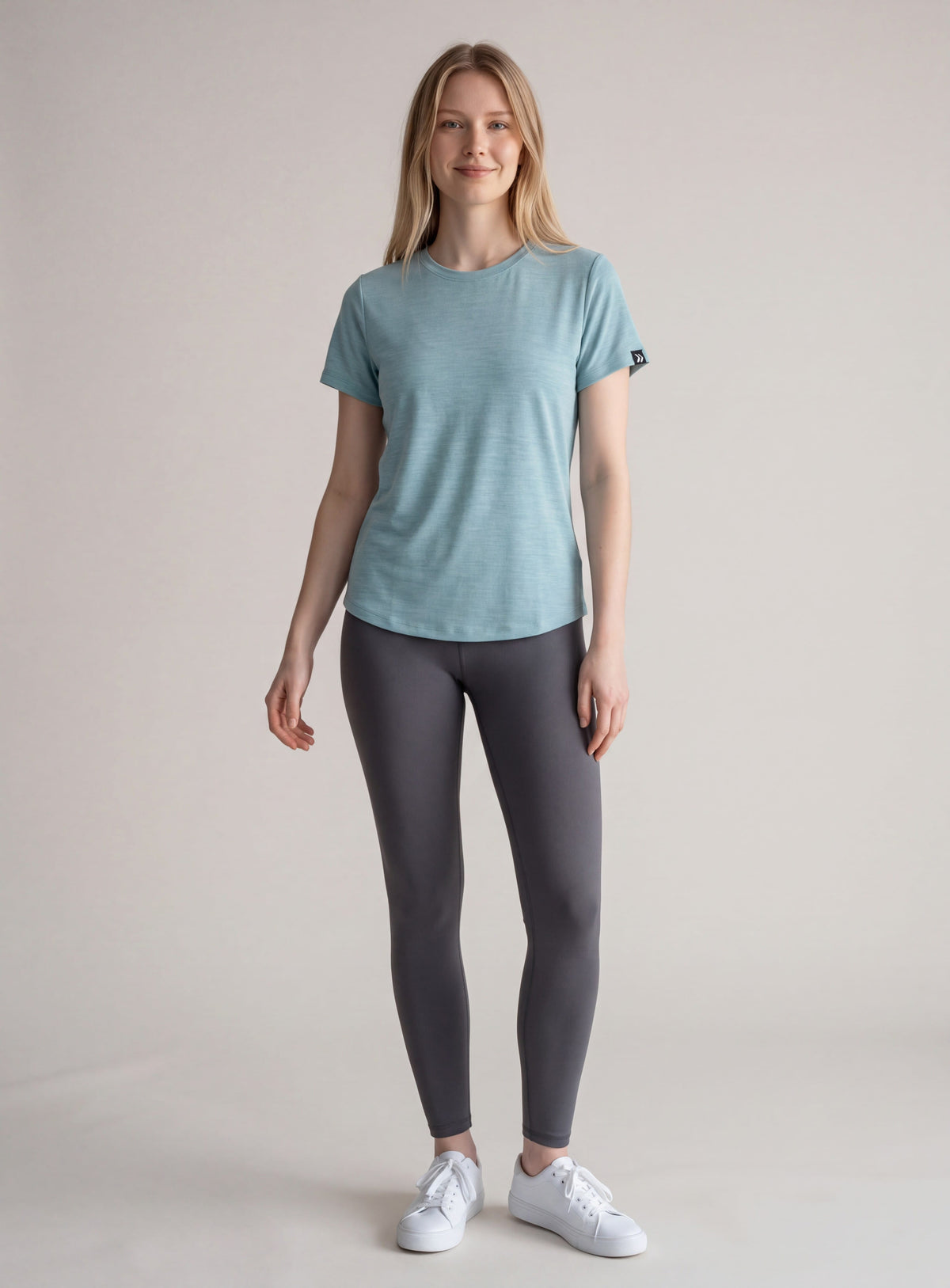 Full-body studio view of a woman wearing MOVE Sport Apparel women’s LuxDry T-shirt in Marina with charcoal leggings and white trainers; original black sleeve tab retained.
