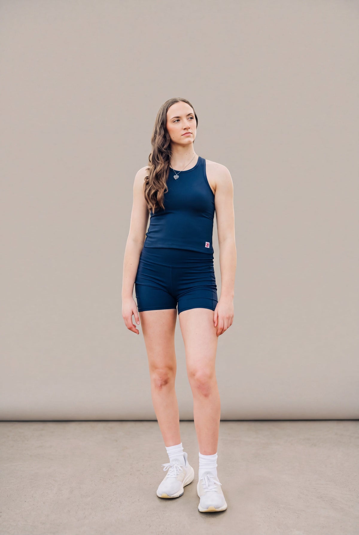 Full-body studio view of a woman wearing the Maritime Blue MOVE Sport Apparel women’s PureStretch Cropped Tank with matching navy shorts and white trainers; original fabric, fit and small red-and-white hem tab retained.