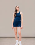 Full-body studio view of a woman wearing the Maritime Blue MOVE Sport Apparel women’s PureStretch Cropped Tank with matching navy shorts and white trainers; original fabric, fit and small red-and-white hem tab retained.