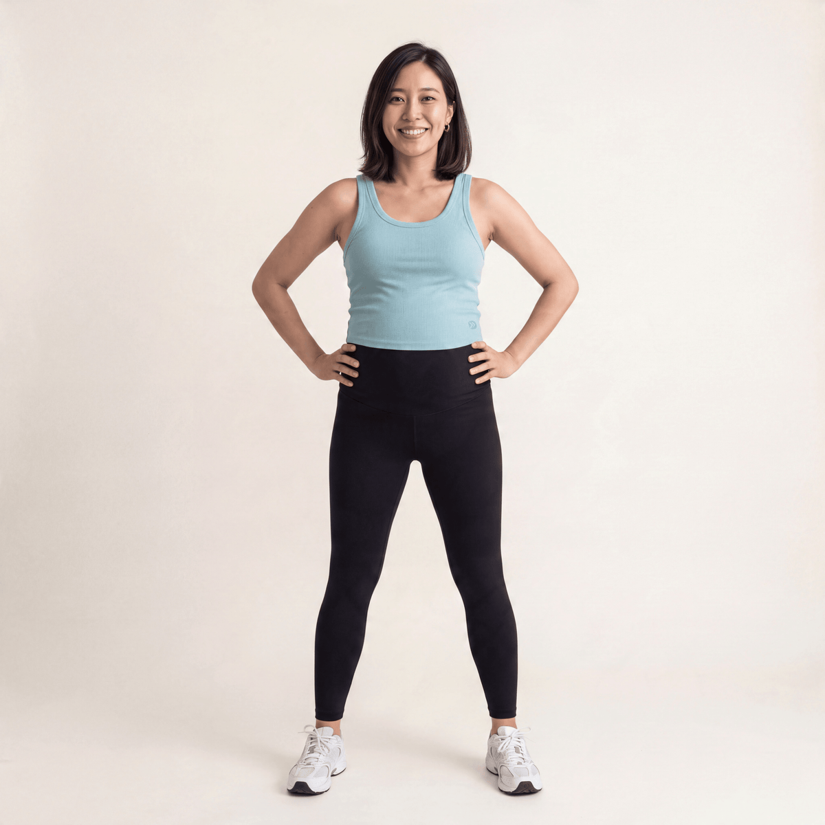 Full-body studio view of a smiling woman wearing the MOVE Sport Apparel Ribbed Bamboo Cropped Tank Top in Aquifer, with black leggings and white trainers.