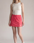 Full-body studio view of a woman wearing MOVE Sport Apparel women’s Skort in Paradise Pink with an ivory sleeveless top and white trainers; original waistband, curved hem and small hem tab retained.