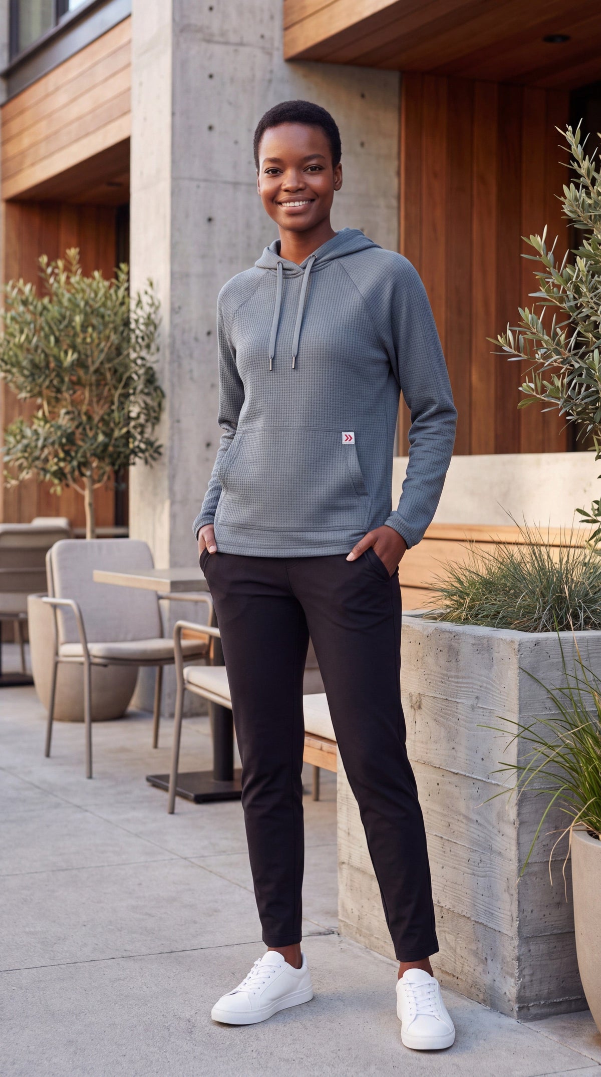Female model wearing MOVE Sport Apparel women’s Balsam Green waffle hoodie beside a modern café terrace, with black pants and white sneakers.