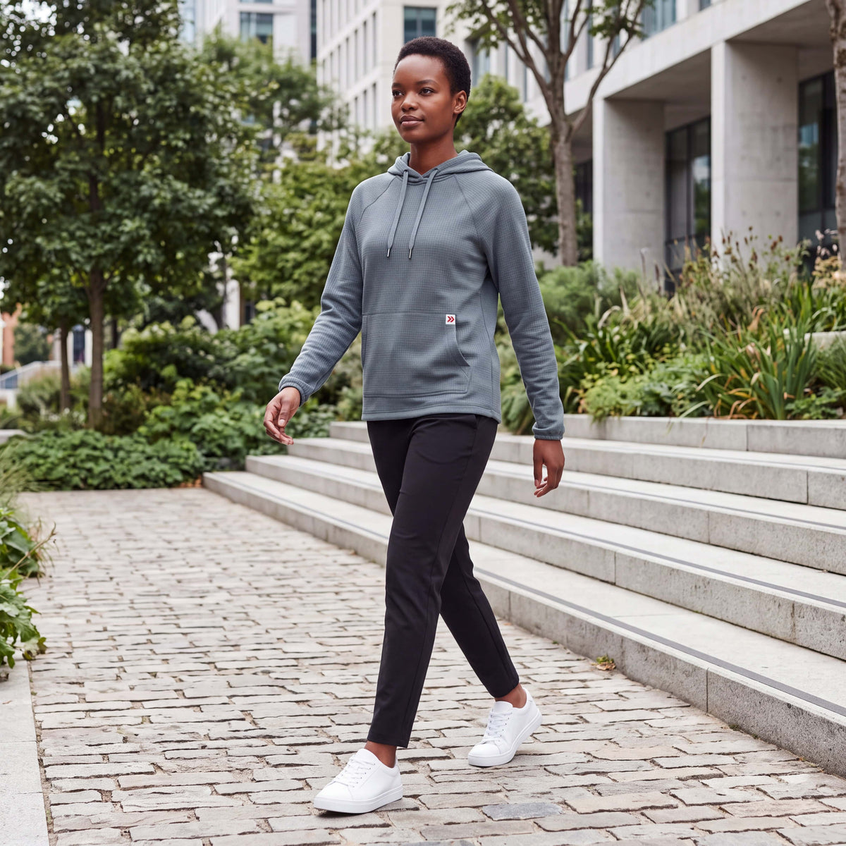 Female model walking through a modern urban park wearing MOVE Sport Apparel women’s Balsam Green waffle hoodie, black training pants and white athletic sneakers.