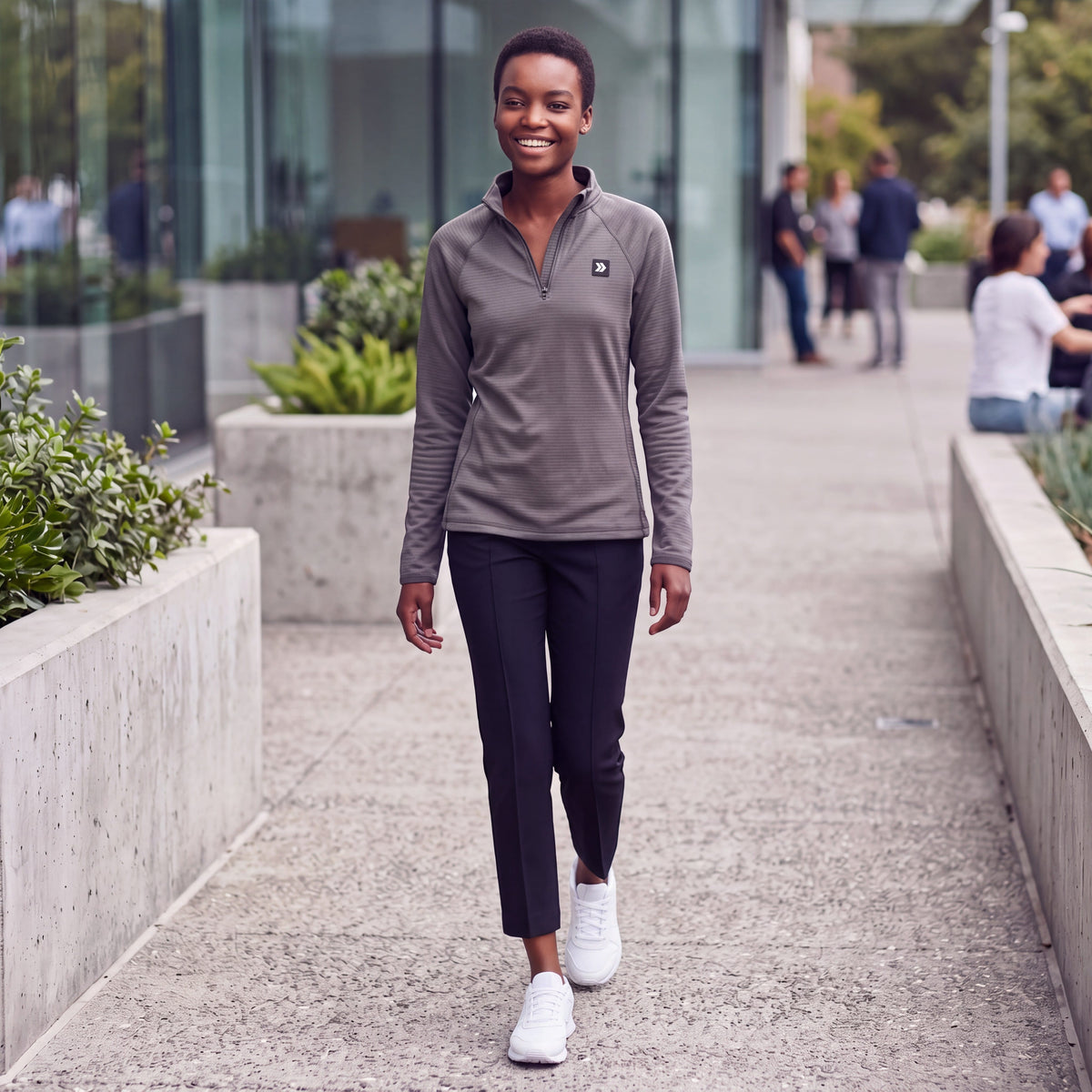 Female model wearing the charcoal MOVE Sport Apparel women’s waffle quarter zip, black pants and white trainers while walking through a modern urban park.