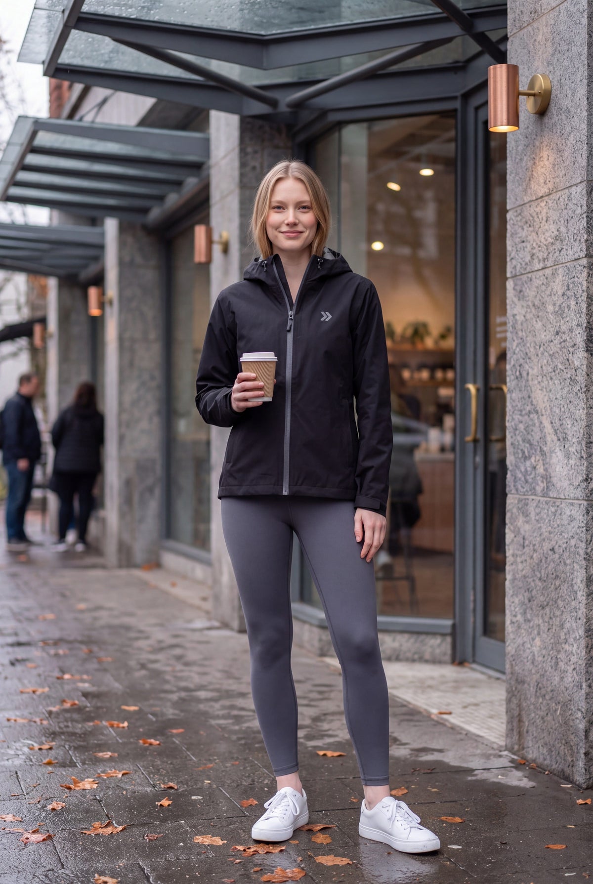 Woman wearing the black MOVE Sport Apparel waterproof jacket outside a café after rain, holding a coffee.