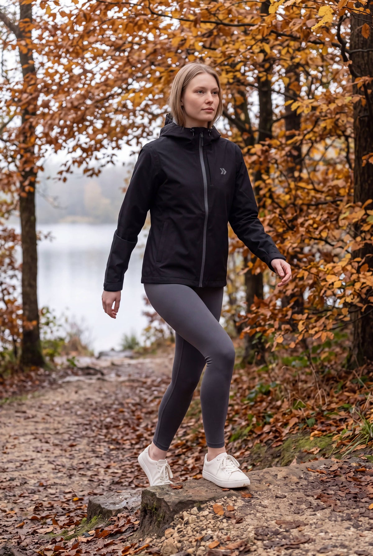 Woman stepping onto a low stone on an autumn woodland trail in the black MOVE Sport Apparel waterproof jacket.